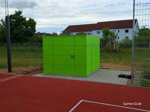 Ein grüner Gartenschrank auf einem Tennisplatz. Im Hintergrund sind Bäume und Gebäude sichtbar.