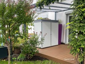 Ein Garten mit einer modernen Holzhütte auf einer Terrasse, umgeben von Pflanzen und einem Tor.