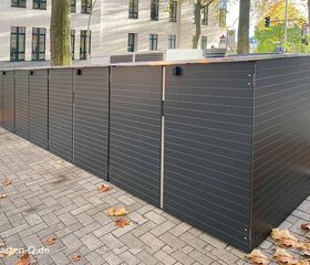 Graue Metallboxen stehen draußen auf einem gepflasterten Bereich neben Bäumen und einem Gebäude.