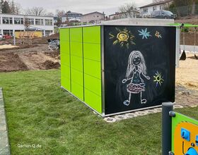 Grüner Schrank mit Tafel bemalt: Kind, Sonne und Sterne in Kreide. Außenbereich mit Rasen und Baustelle.