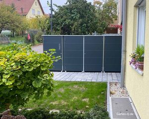 Ein großer, schwarzer Paketkasten im Garten neben einem Haus, umgeben von Pflanzen und Gehwegplatten.