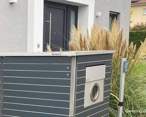 Ein moderner Eingang mit Pflanzen, einem dunkelgrauen Zaun und einer runden Metallöffnung.