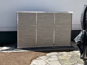 Ein grauer Gartenschrank steht auf einer gepflasterten Fläche neben einer Mauer.