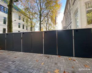 Moderne, dunkle Gartenboxen stehen auf einem gepflasterten Weg vor einem weißen Gebäude, umgeben von Bäumen im Herbst.