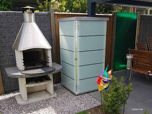 . Außenkamin und Gartenbox neben Gartenmöbeln und farbigem Windrad.