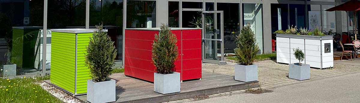 Pflanzen in Töpfen vor einem Gebäude mit bunten Kuben und Glasfront, sonniger Tag.